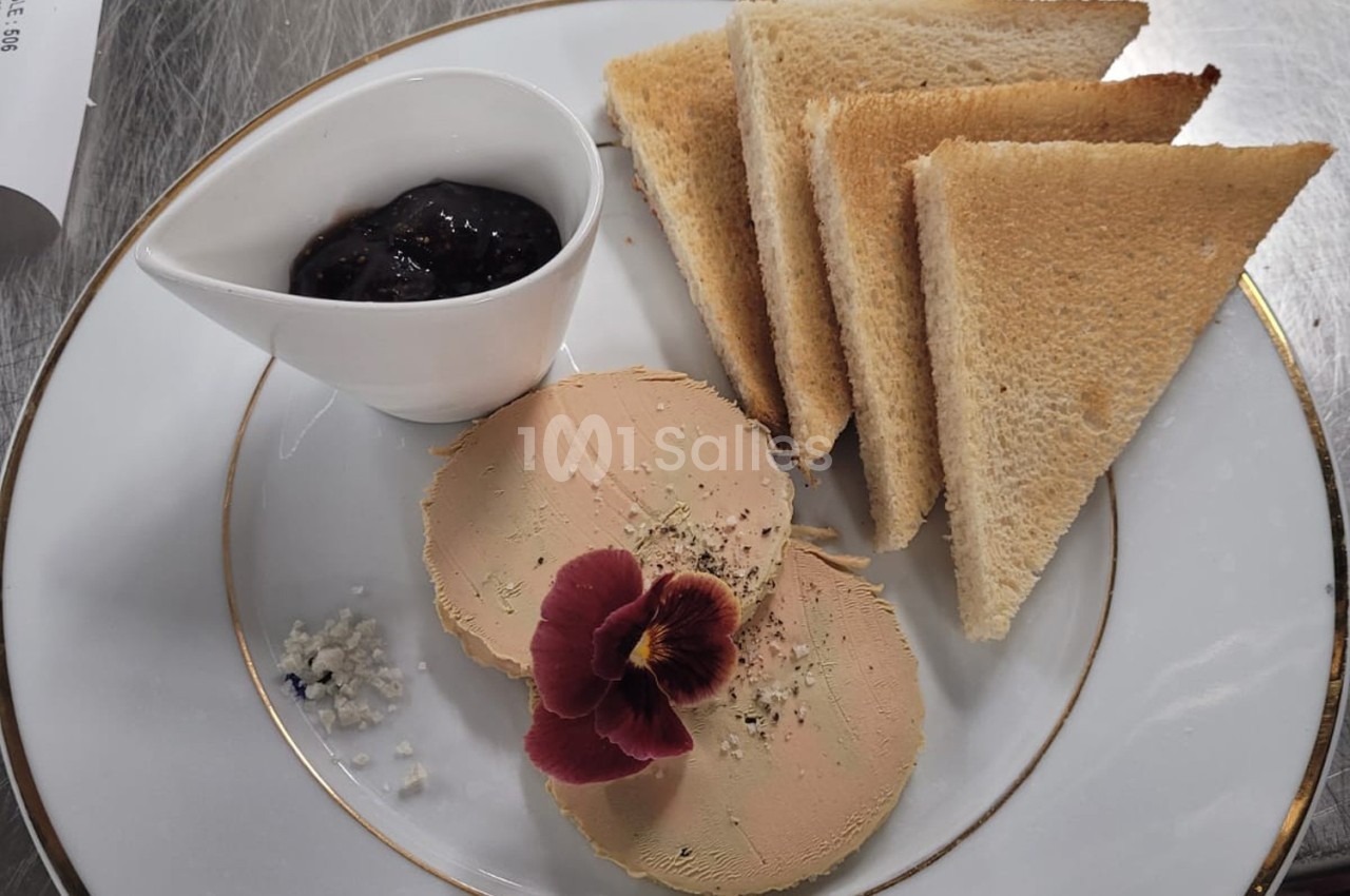 Assiette avec deux tranches de foie gras, une fleur comestible, du sel, des toasts triangulaires et un ramequin de confiture.