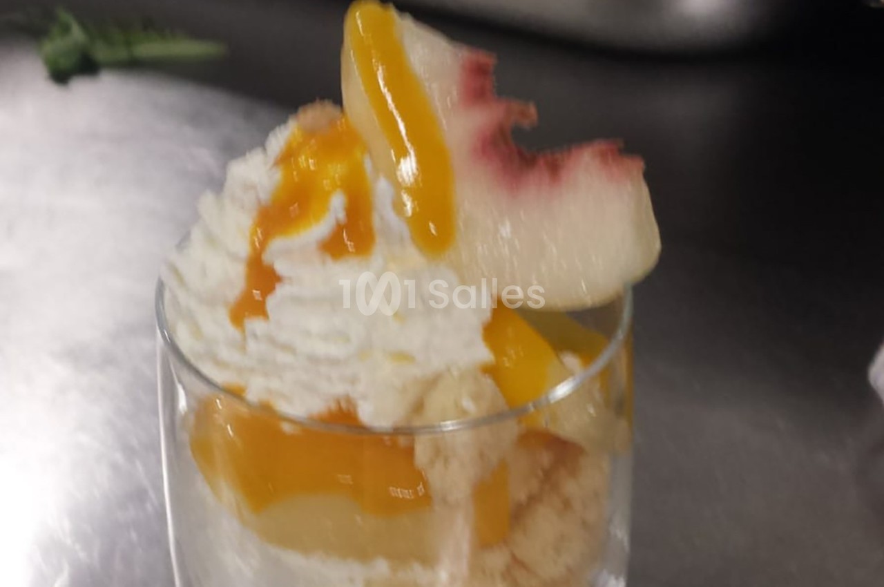 Verrine de dessert composée de crème fouettée, morceaux de pêche et coulis de mangue sur un lit de biscuit.