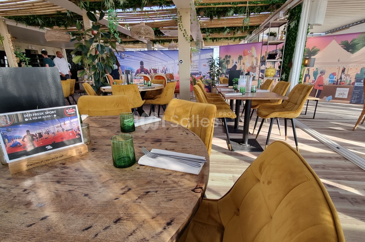 Salle de restaurant lumineuse avec tables en bois, chaises jaunes, décoration végétale et fresques colorées en arrière-plan.