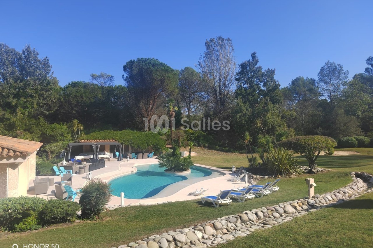 Piscine extérieure entourée de transats, pelouse et arbres, avec une terrasse aménagée sous un ciel dégagé.