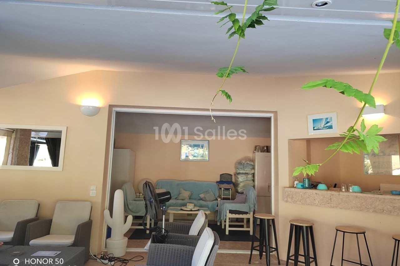 Intérieur d'une pièce lumineuse avec salon, bar, tabourets et plantes suspendues au premier plan.