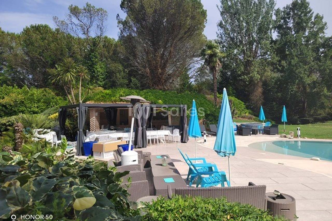 Terrasse aménagée avec mobilier de jardin, parasols bleus et piscine, entourée de végétation dense et arbres.