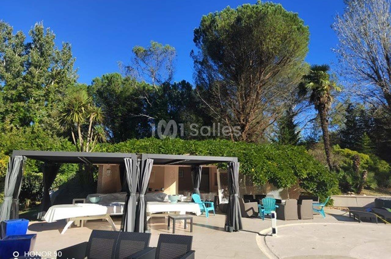 Espace extérieur avec pergolas, tables et chaises, entouré de végétation et arbres sous un ciel bleu clair.