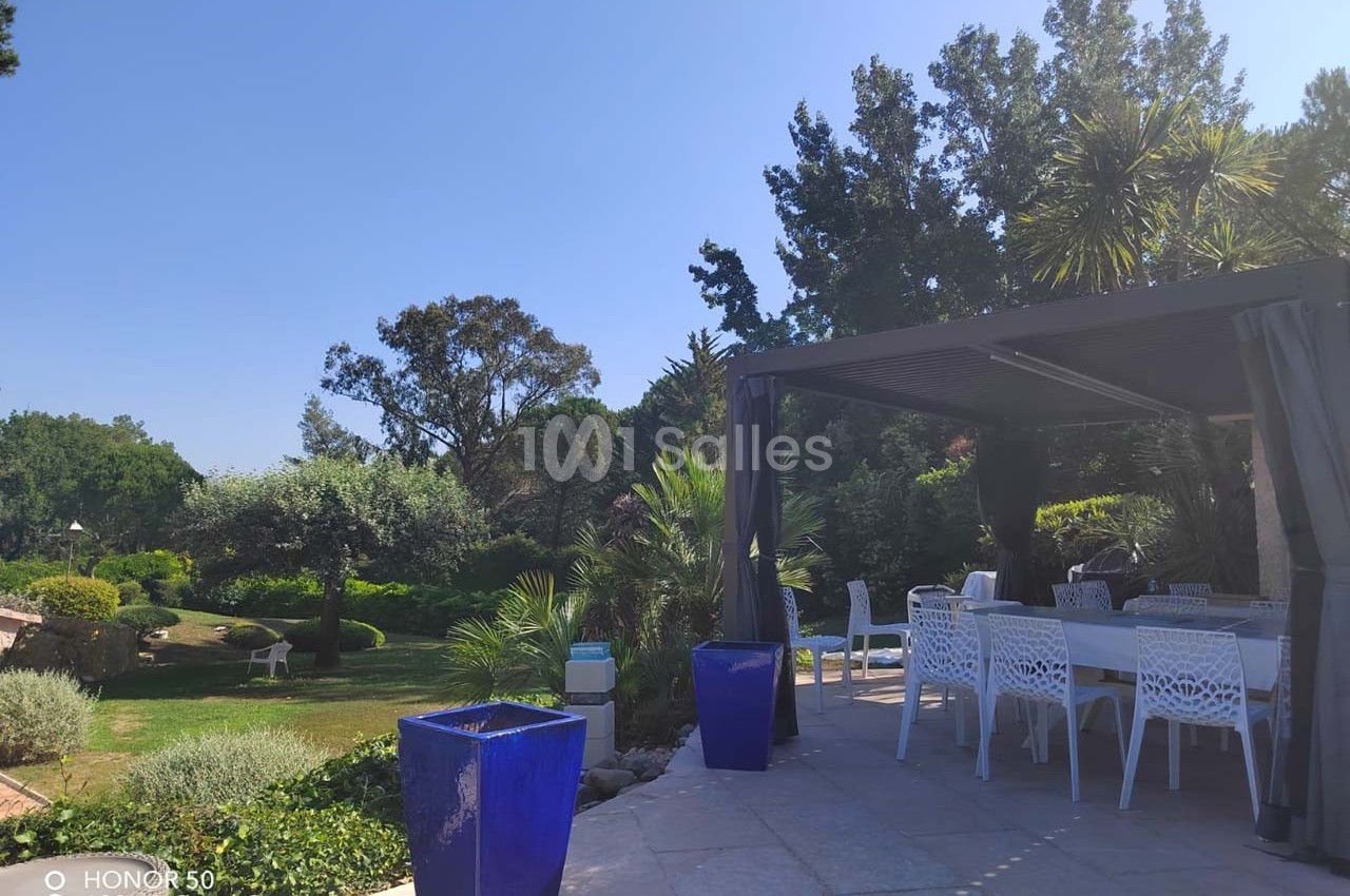 Terrasse avec table et chaises blanches sous une pergola, entourée de plantes et arbres dans un jardin ensoleillé.