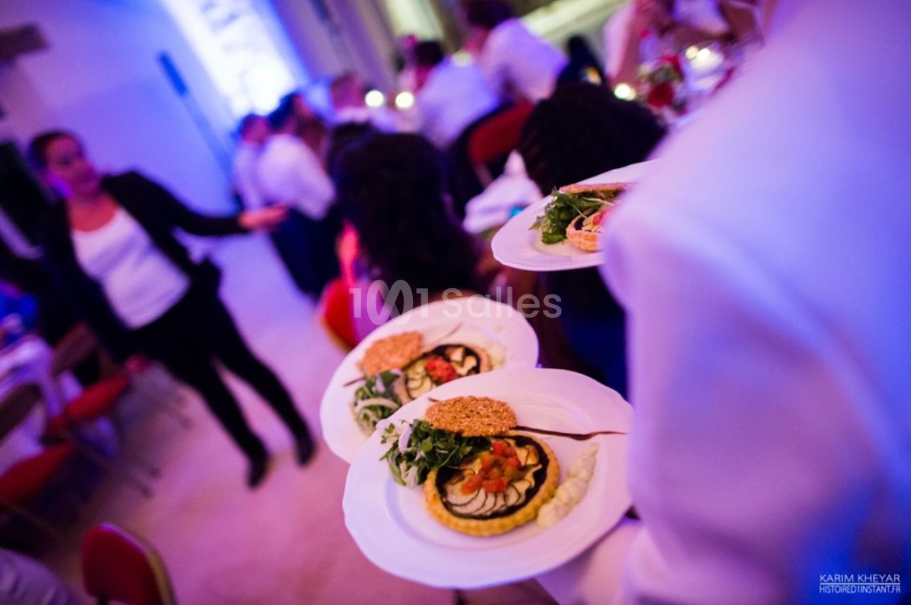 Serveur apportant plusieurs assiettes garnies de tartes salées, salades et crackers dans une salle de réception animée.
