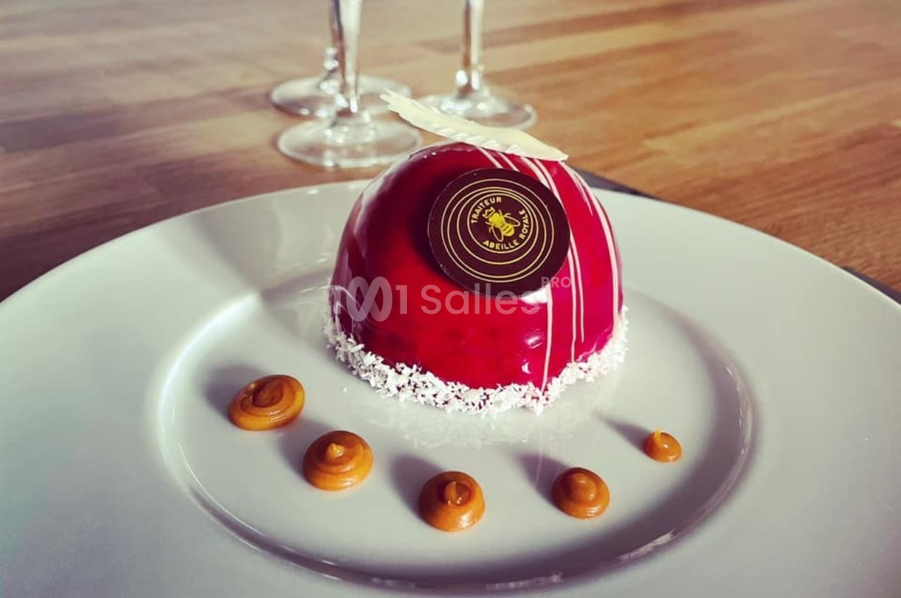 Entremets rouge brillant sur assiette blanche, décoré de caramel, noix de coco râpée et chocolat, avec des verres en arrière…