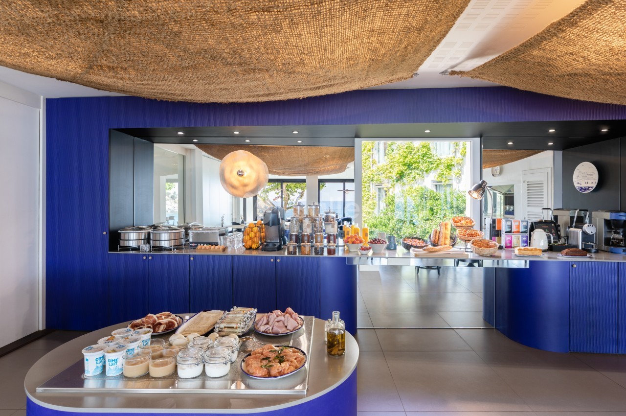 Buffet de petit-déjeuner avec viennoiseries, charcuteries, produits laitiers et fruits, dans une salle lumineuse.