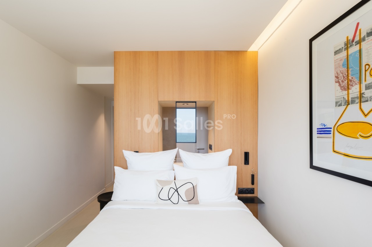 Lit double avec oreillers blancs dans une chambre moderne, décorée de bois clair et d'un tableau encadré.