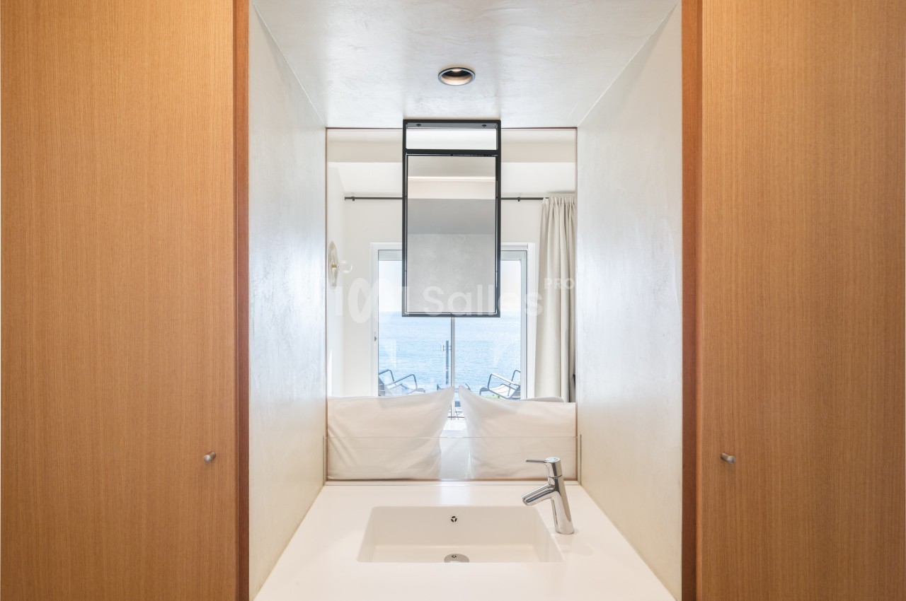Lavabo blanc avec robinet moderne devant un miroir reflétant une vue sur la mer et des chaises sur un balcon.