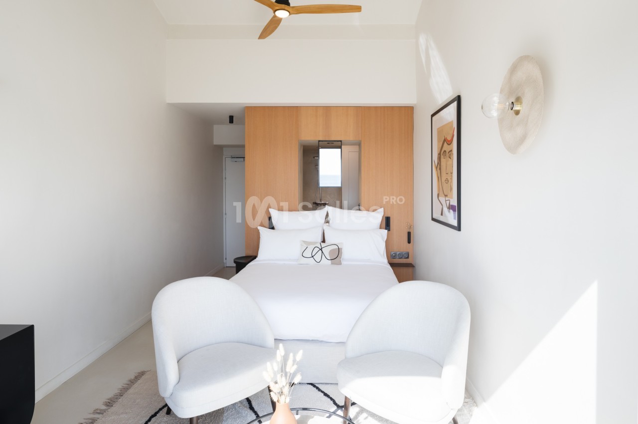 Chambre lumineuse avec lit double, deux fauteuils blancs, mur en bois clair et décoration minimaliste.