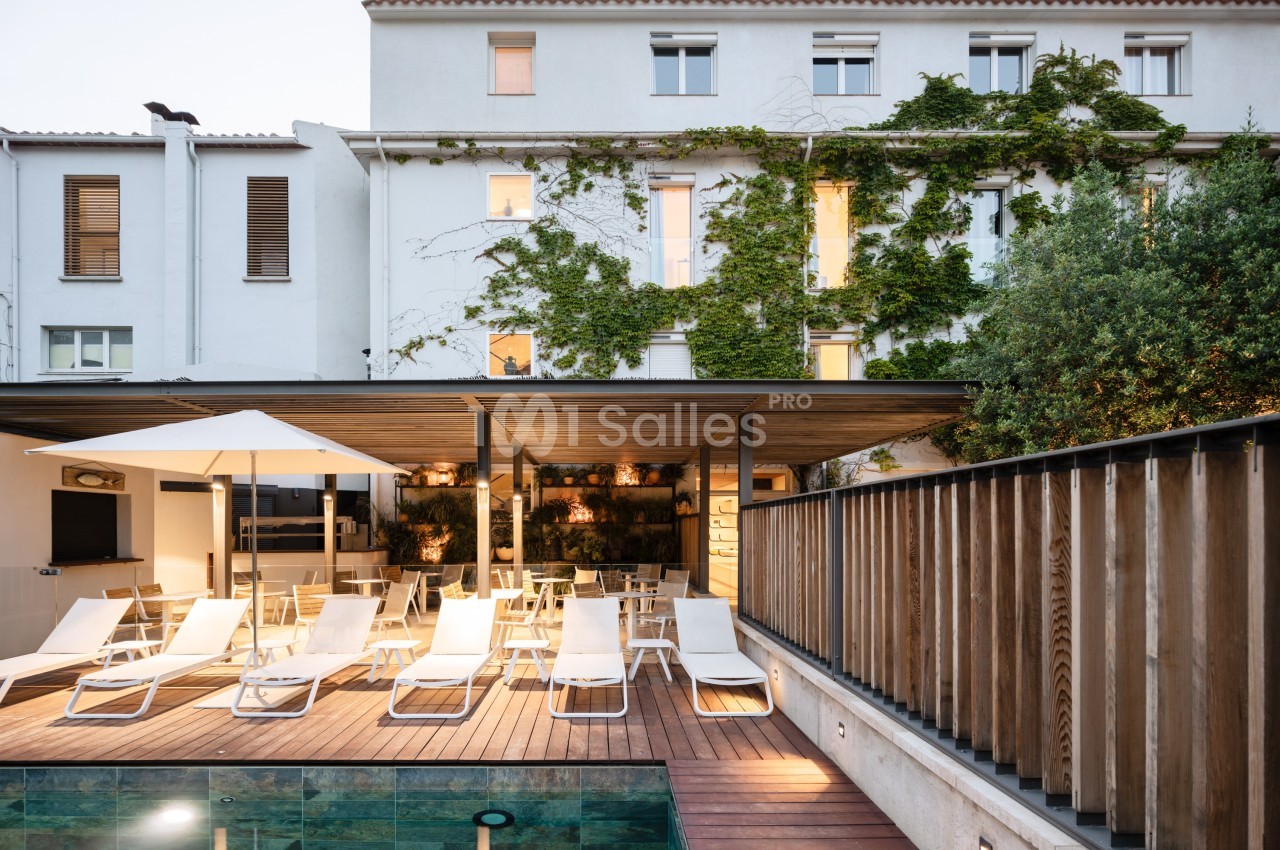 Façade d'un bâtiment avec terrasse en bois, chaises longues, piscine et végétation grimpante sur les murs.