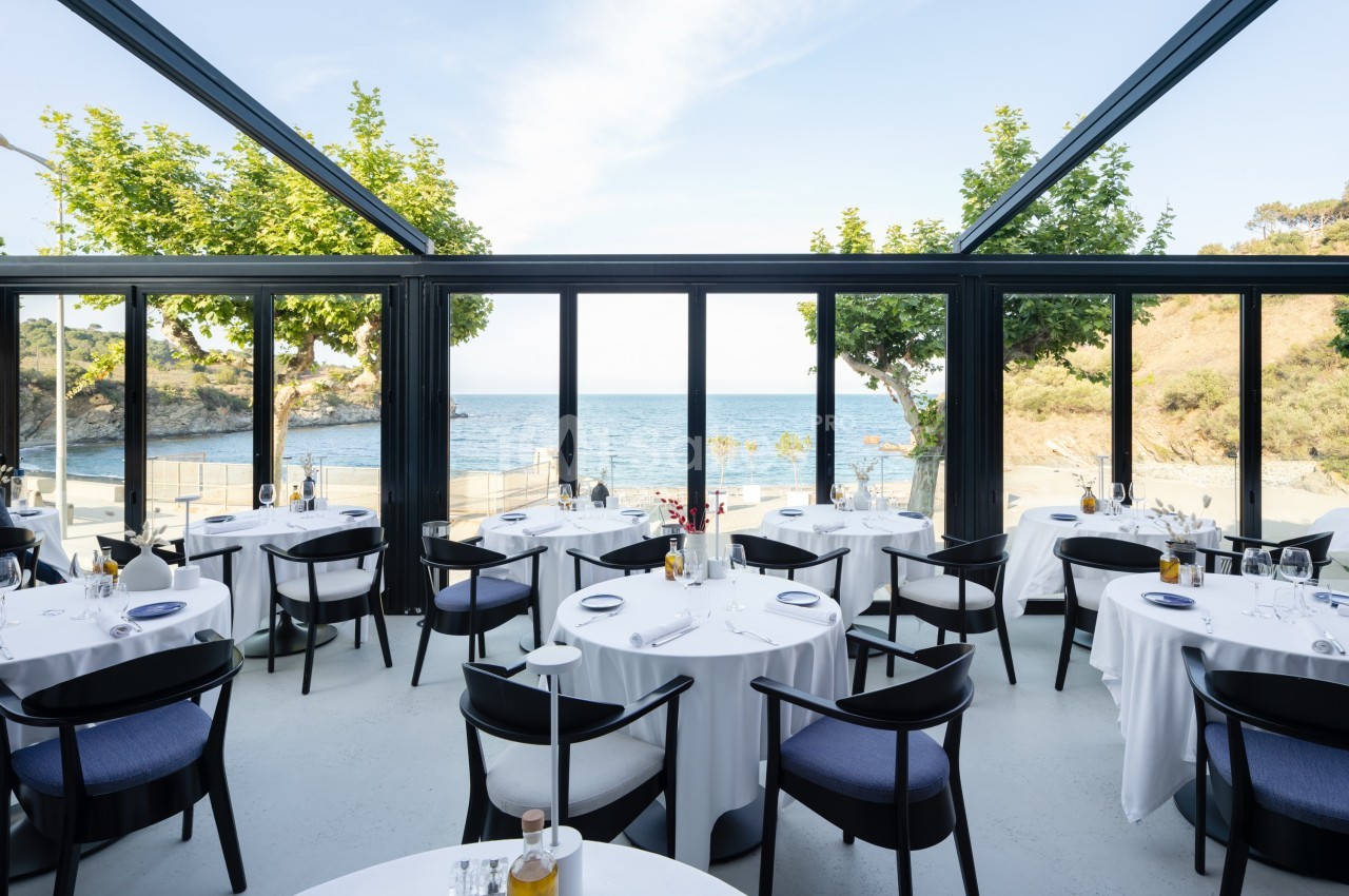 Salle de restaurant lumineuse avec baies vitrées donnant sur une vue dégagée sur la mer et des collines.