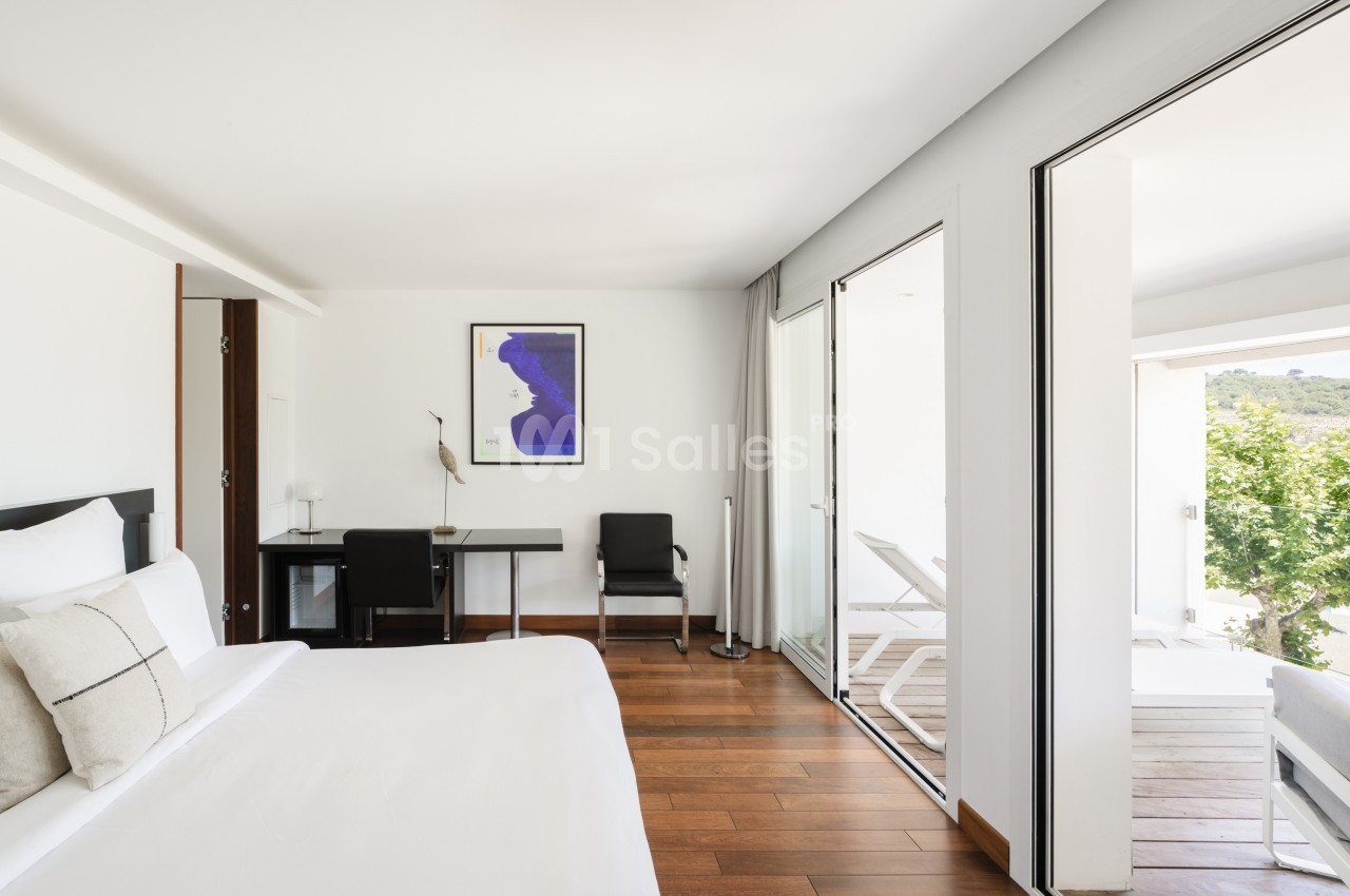 Chambre lumineuse avec lit double, bureau, chaises, et accès à une terrasse avec vue sur la végétation.