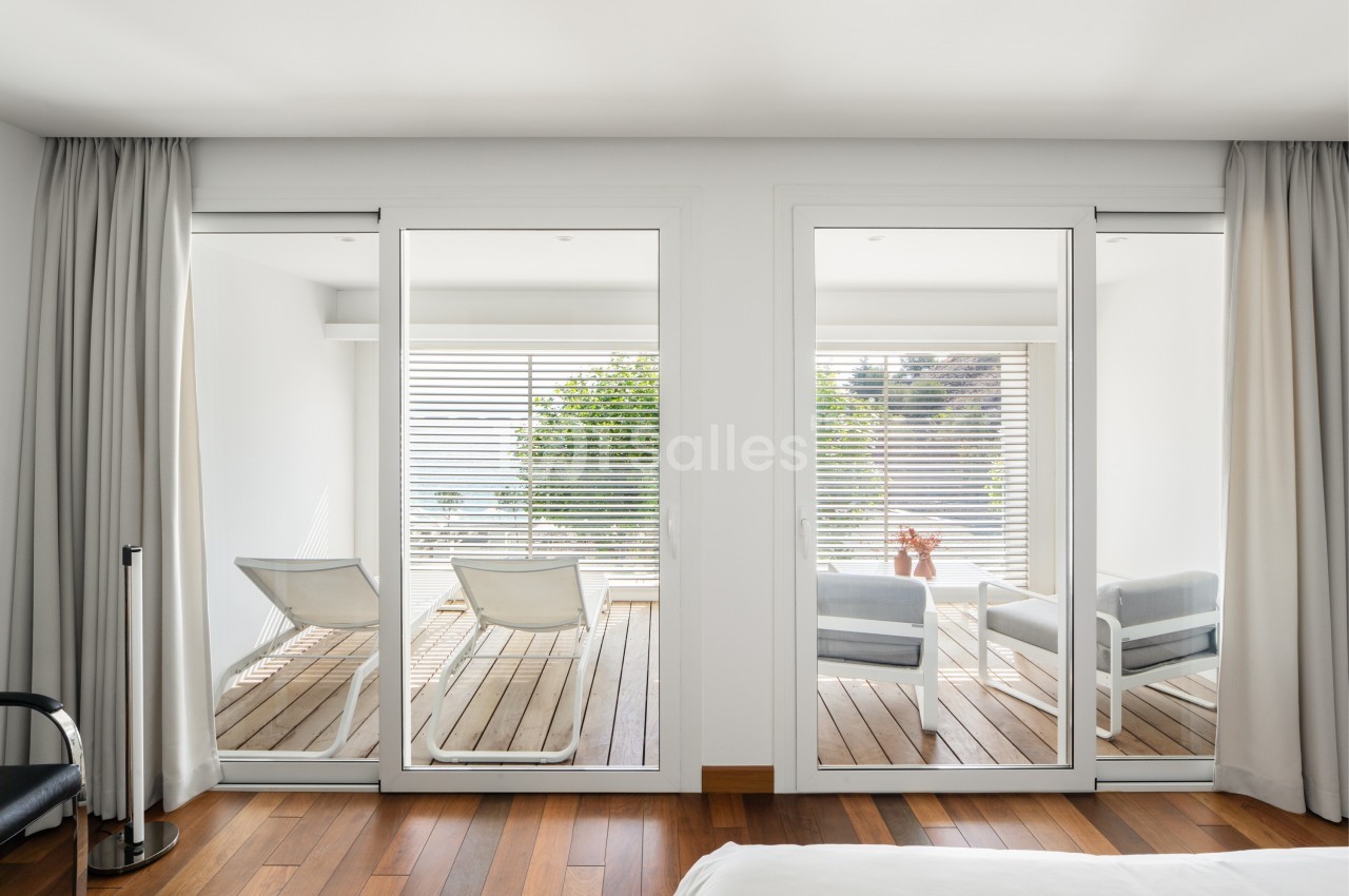 Vue depuis une chambre sur une terrasse en bois avec deux chaises longues et un fauteuil, entourée de baies vitrées.