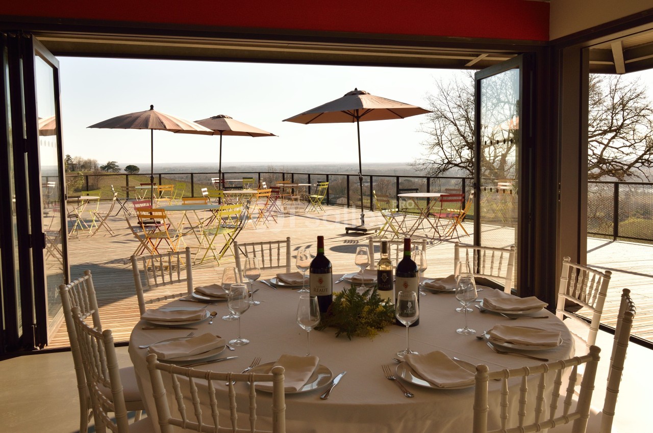 Terrasse ensoleillée avec tables et chaises colorées, vue dégagée, table dressée à l'intérieur avec bouteilles de vin.