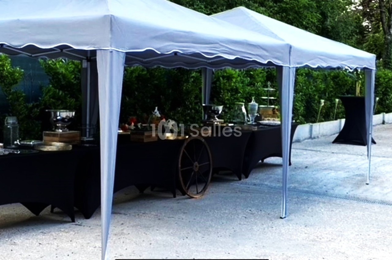 Tentes blanches abritant des tables noires dressées pour un buffet en extérieur, entourées de verdure.