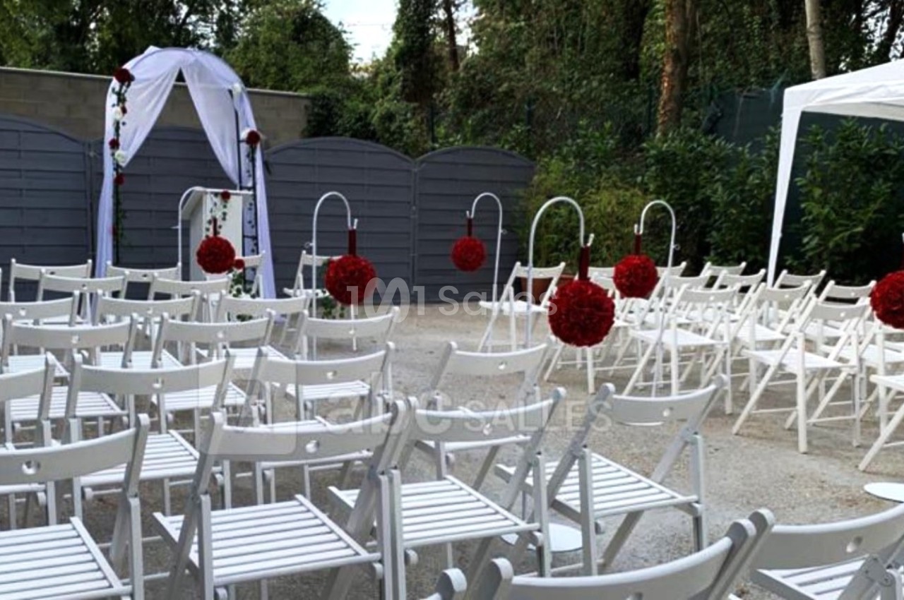 Chaises blanches disposées en rangées pour une cérémonie en extérieur, avec une arche décorée et des fleurs rouges.
