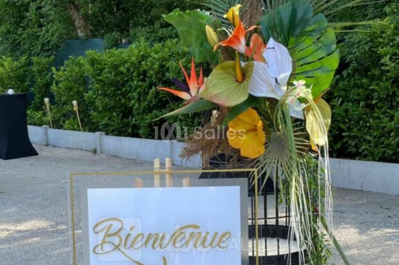 Panneau ’Bienvenue’ décoré de fleurs tropicales colorées, placé à l'extérieur près d'une haie et de tables hautes noires.