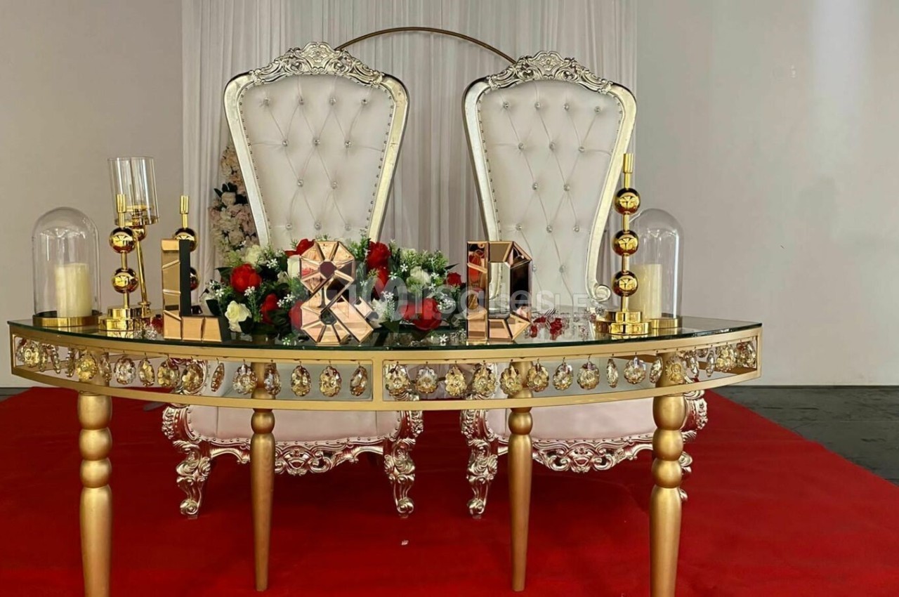 Table décorative dorée avec ornements floraux et bougies, devant deux fauteuils blancs capitonnés sur tapis rouge.