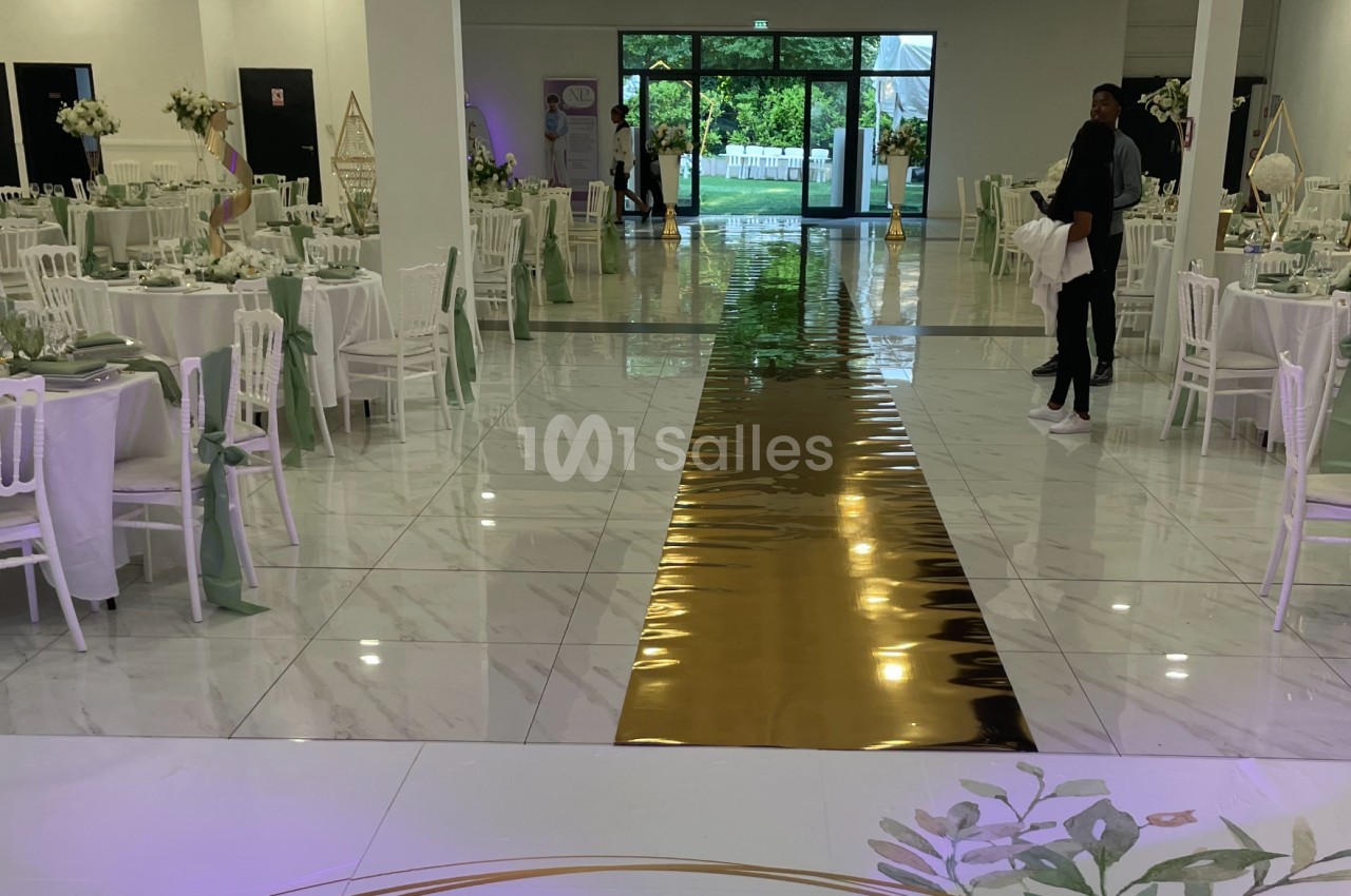Salle de réception décorée avec des tables élégantes, un chemin doré central et une vue sur un jardin à travers les baies…