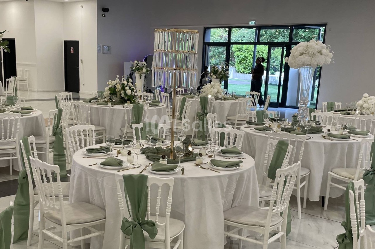 Salle de réception décorée avec des tables rondes, nappes blanches, chaises ornées de rubans verts et compositions florales.