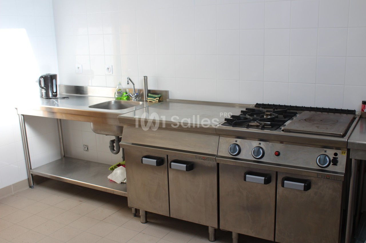 Cuisine professionnelle avec plan de travail en inox, évier, bouilloire, plaques de cuisson et carrelage mural blanc.