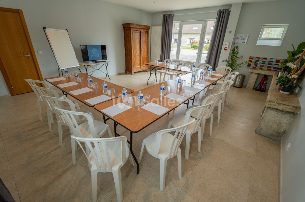 Salle de réunion lumineuse avec tables disposées en U, chaises en plastique, bouteilles d'eau et écran de présentation.