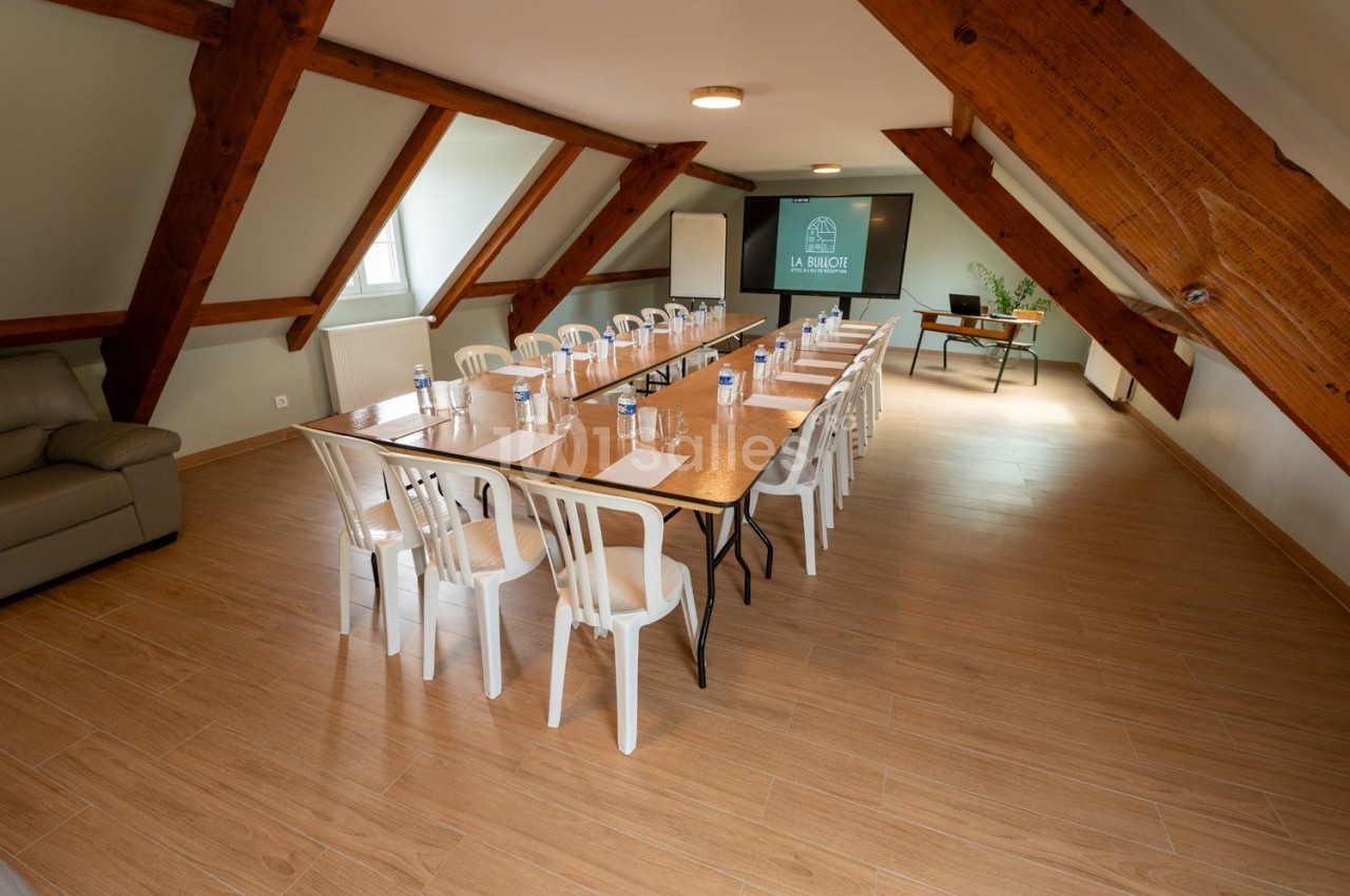 Salle de réunion sous combles avec grande table, chaises blanches, bouteilles d'eau, blocs-notes et écran de présentation.