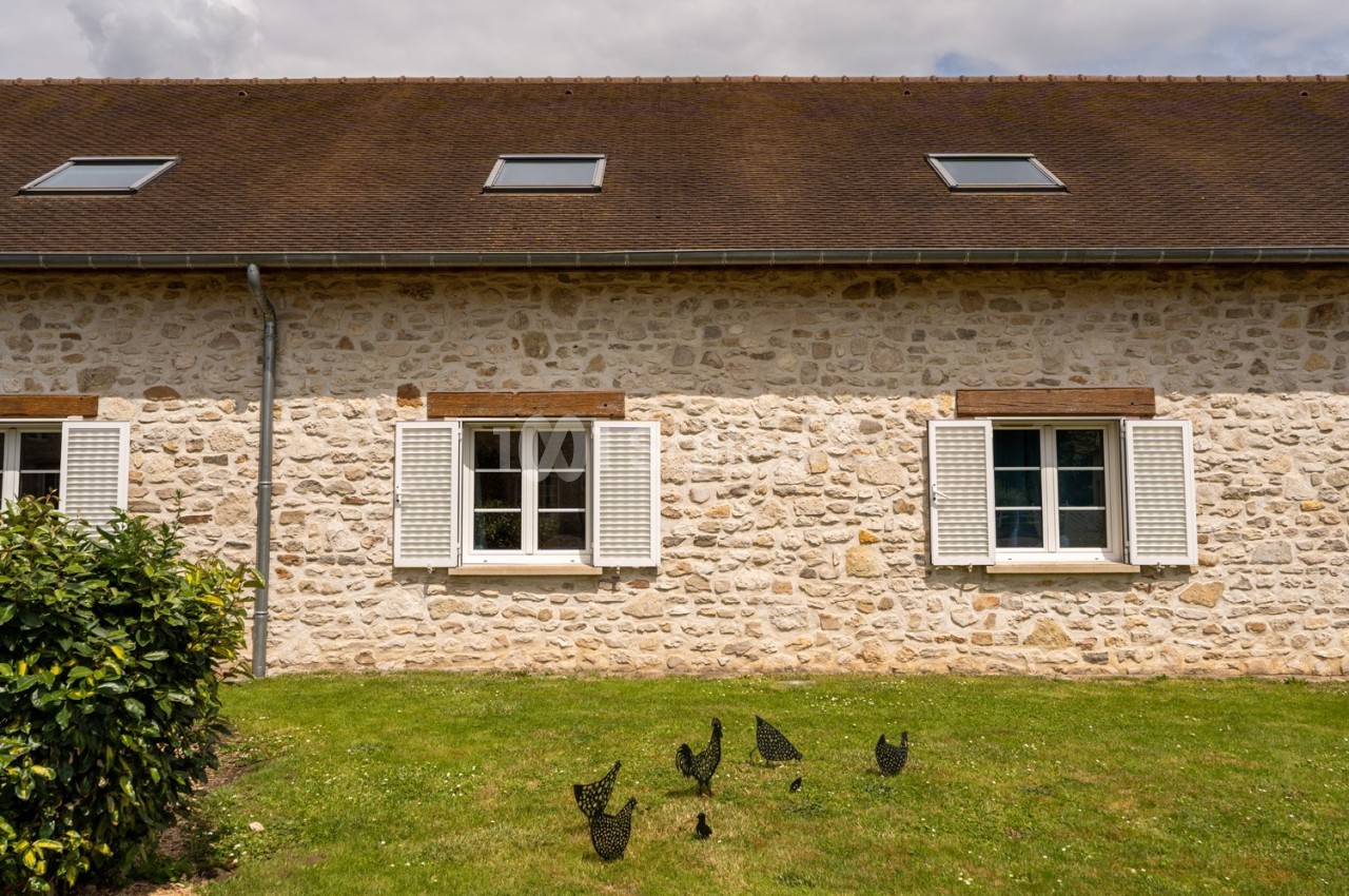 Façade en pierre d'une maison avec volets blancs, fenêtres mansardées et pelouse ornée de sculptures métalliques.