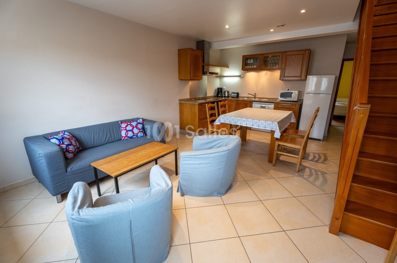 Salon avec canapé, fauteuils, table basse et cuisine ouverte équipée, dans un intérieur lumineux avec carrelage beige.