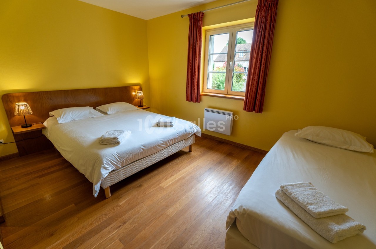 Chambre lumineuse avec murs jaunes, lit double, lit simple, parquet en bois et fenêtre avec rideaux rouges.