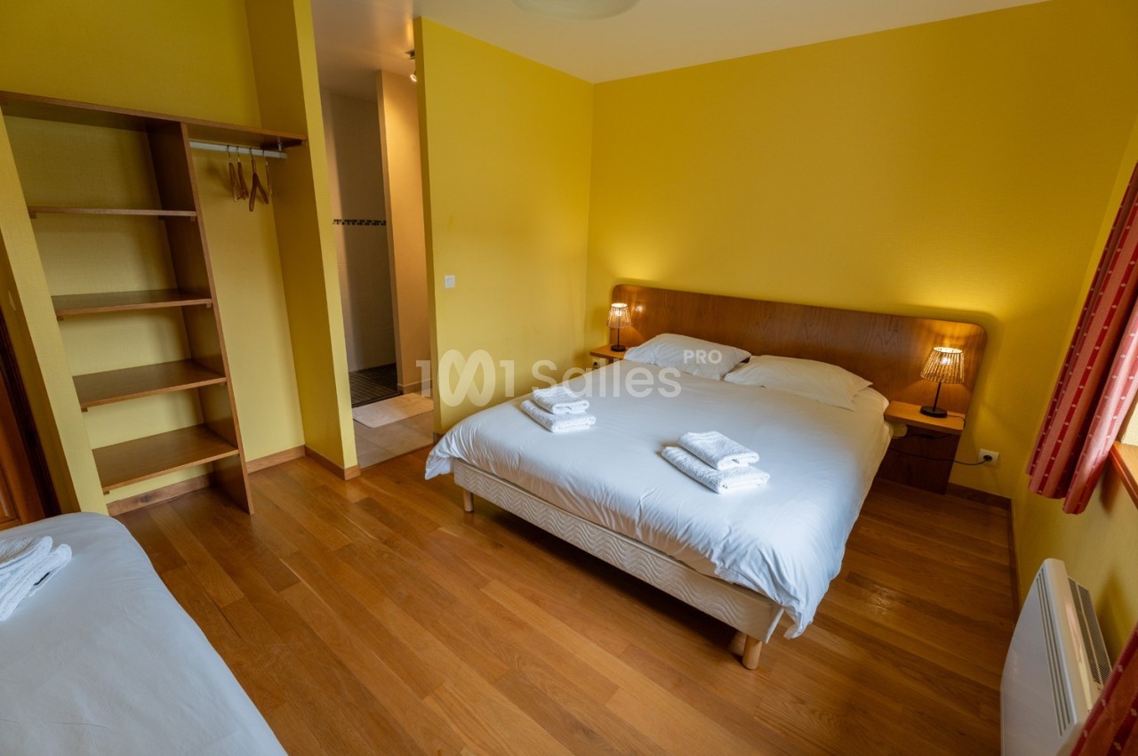 Chambre avec lit double, lit simple, parquet, murs jaunes, étagères ouvertes et salle de bain attenante visible.