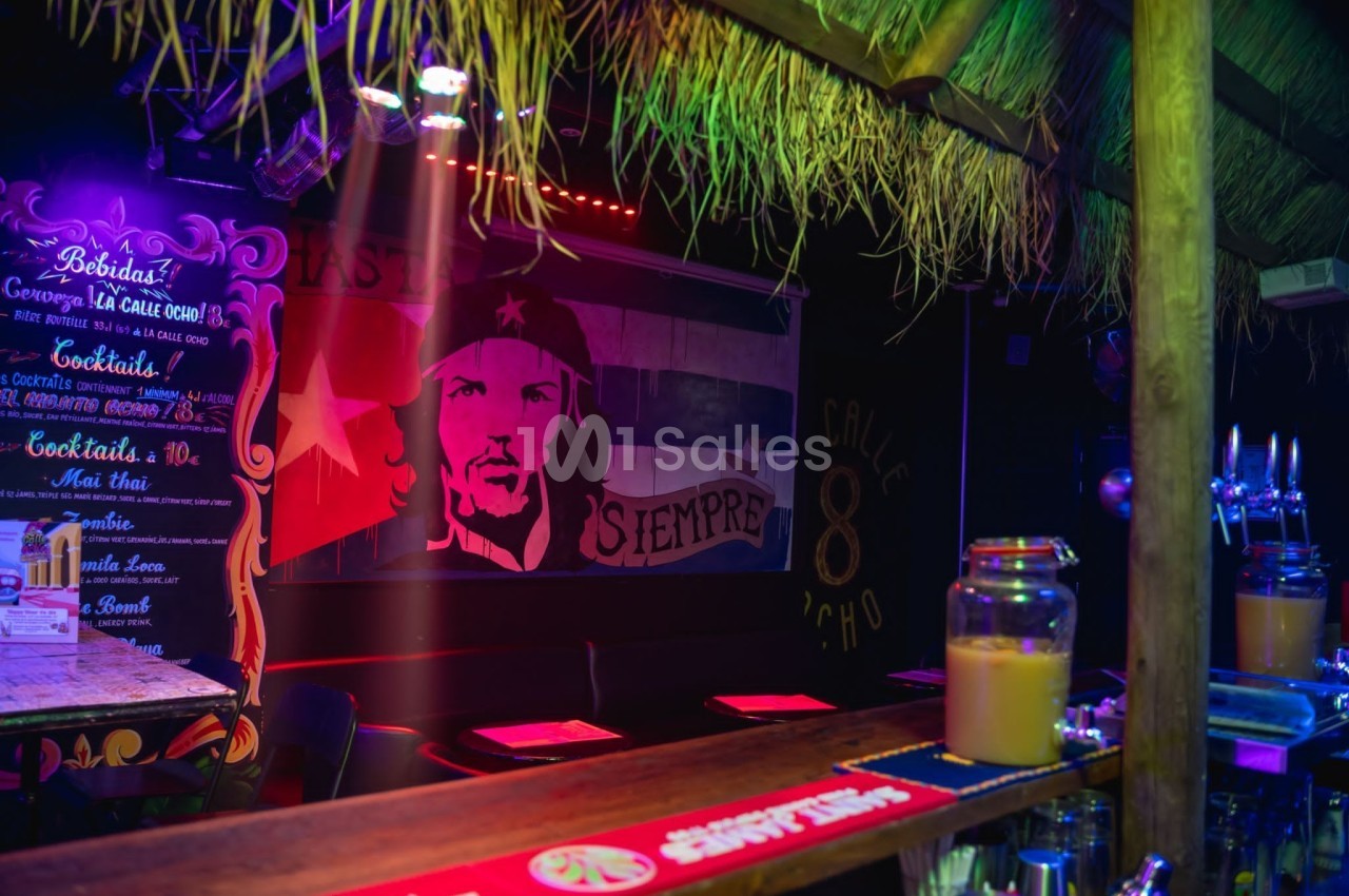 Intérieur d'un bar coloré avec un décor tropical, un portrait stylisé sur un mur et un comptoir avec des boissons.