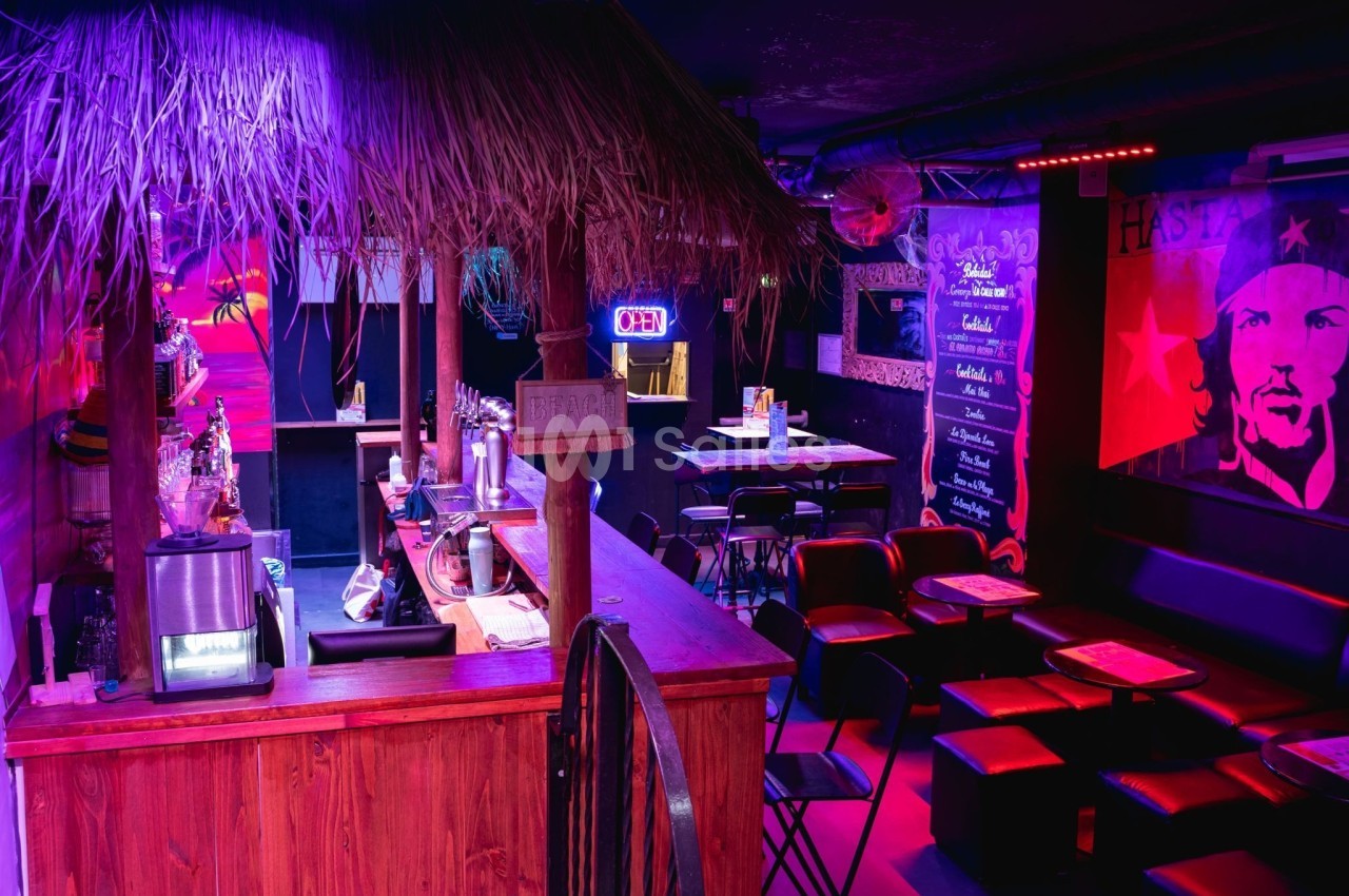 Bar avec comptoir en bois, chaises noires, éclairage coloré et décoration murale à thème tropical et révolutionnaire.