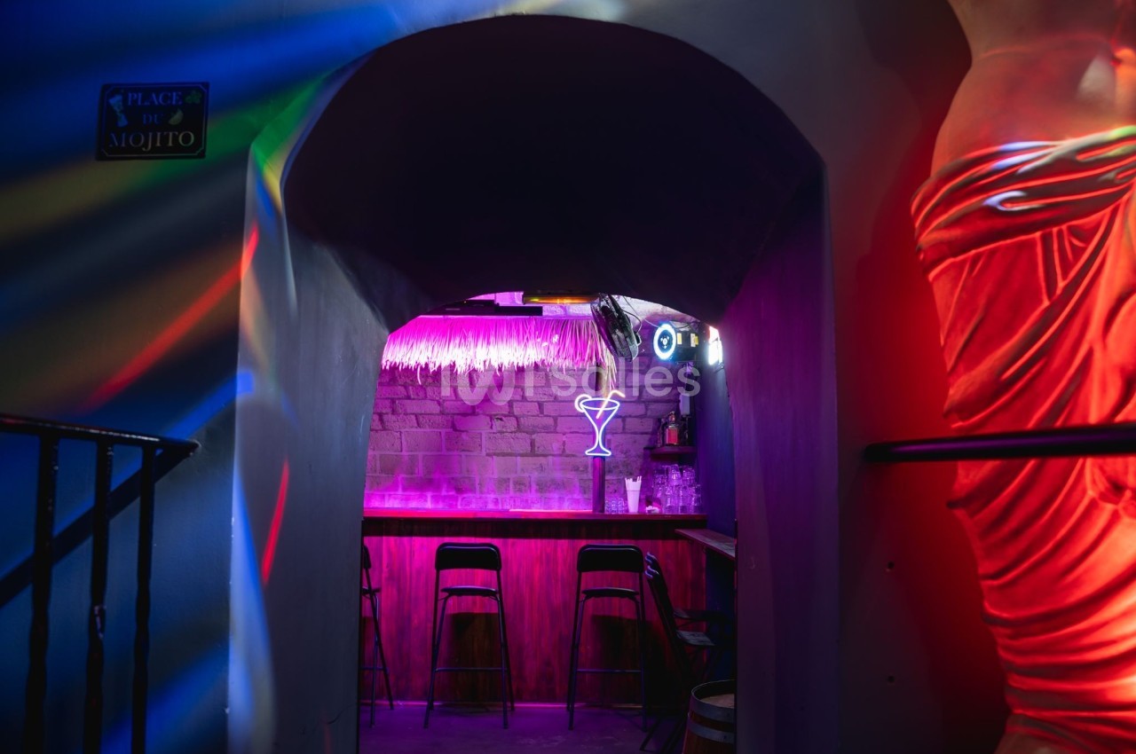 Bar coloré avec tabourets, éclairage néon violet et enseigne en forme de verre à cocktail dans une ambiance tamisée.