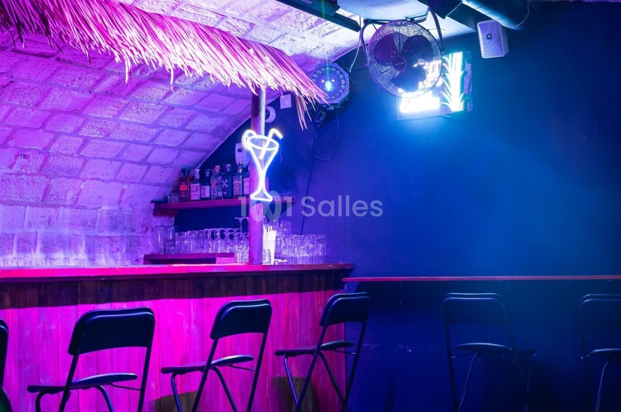 Bar avec comptoir en bois, tabourets alignés, éclairage violet et néon en forme de verre dans une ambiance tamisée.