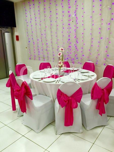 Table ronde dressée avec nappes blanches, chaises décorées de nœuds roses et centre de table floral, dans une salle ornée de…