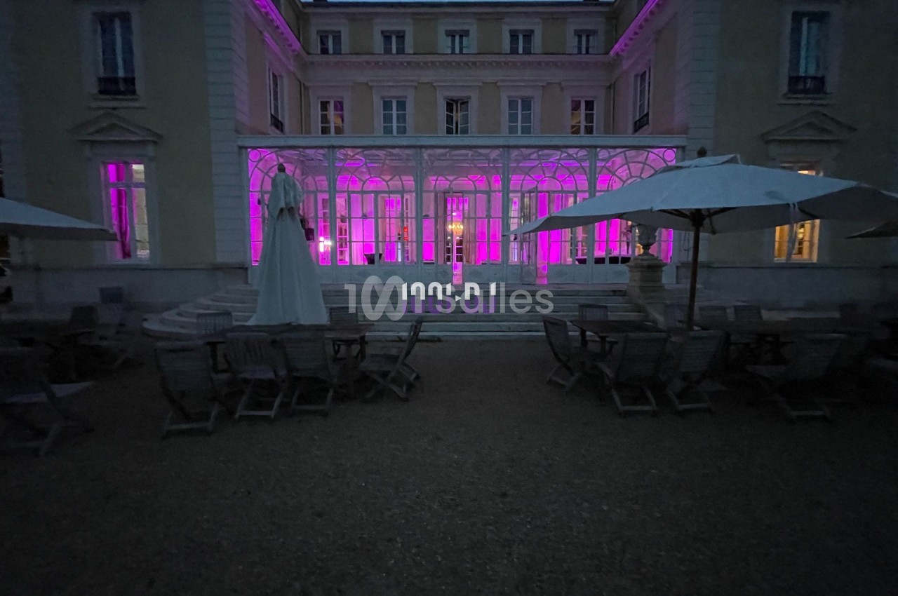 Façade d'un bâtiment éclairé en rose, avec terrasse aménagée de tables et parasols au premier plan, en soirée.