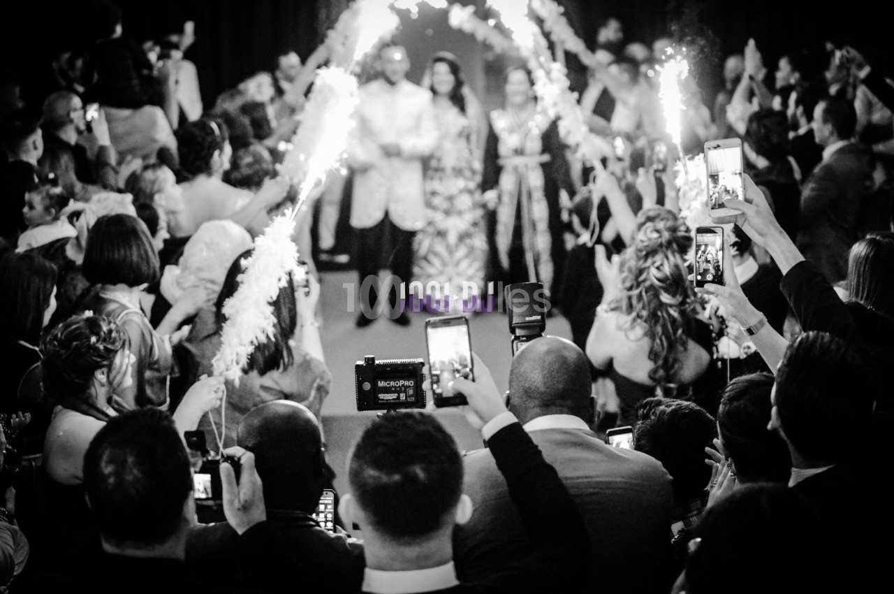 Des mariés avancent sous des étincelles tenues par des invités, entourés d'une foule prenant des photos.