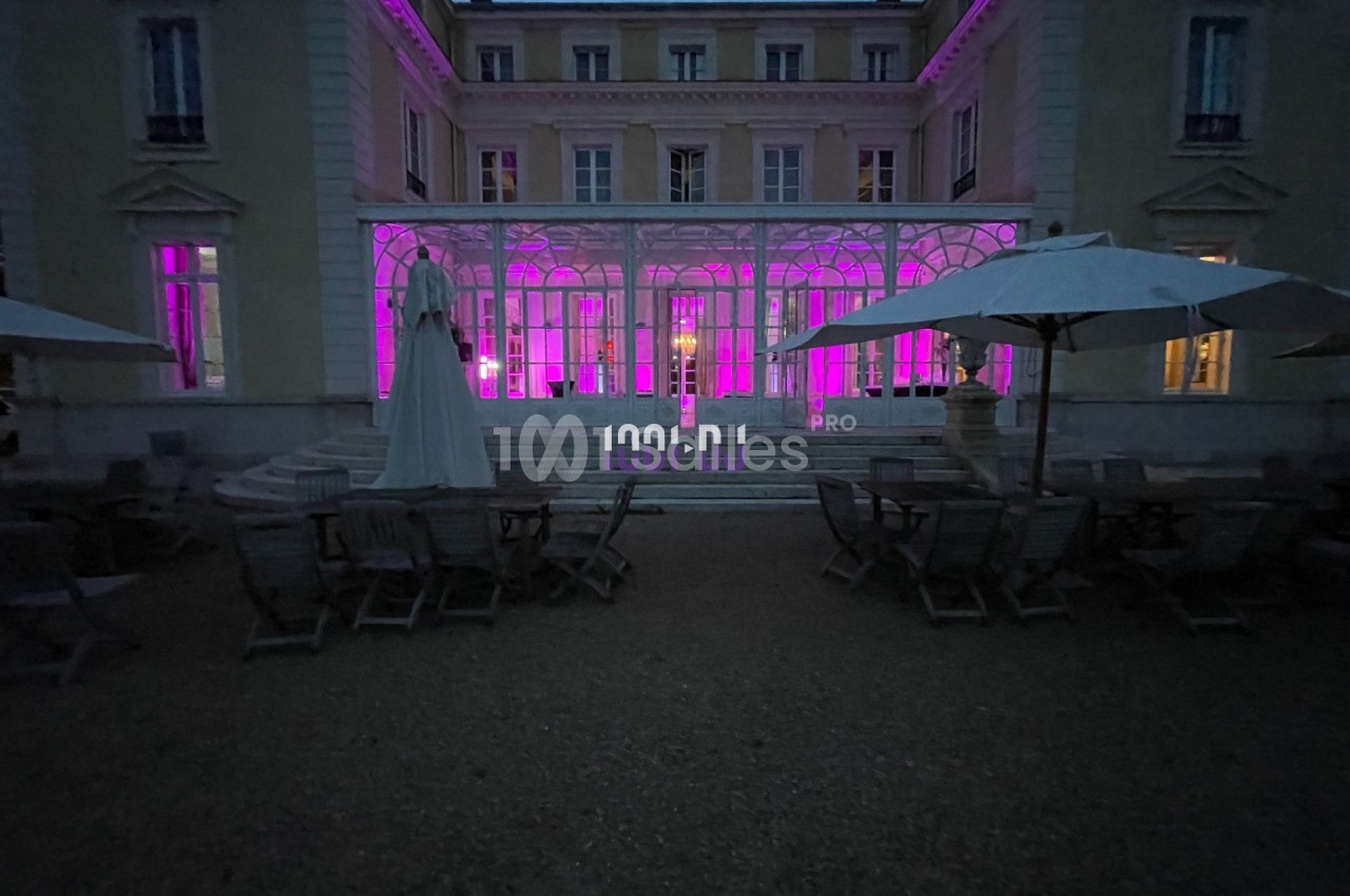 Façade d'un bâtiment éclairé en rose, avec terrasse aménagée de tables et parasols au premier plan, en soirée.