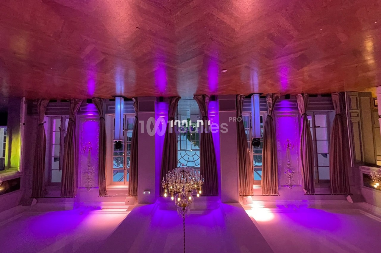 Salle élégante avec parquet, lustre central et éclairage violet mettant en valeur des fenêtres avec rideaux.
