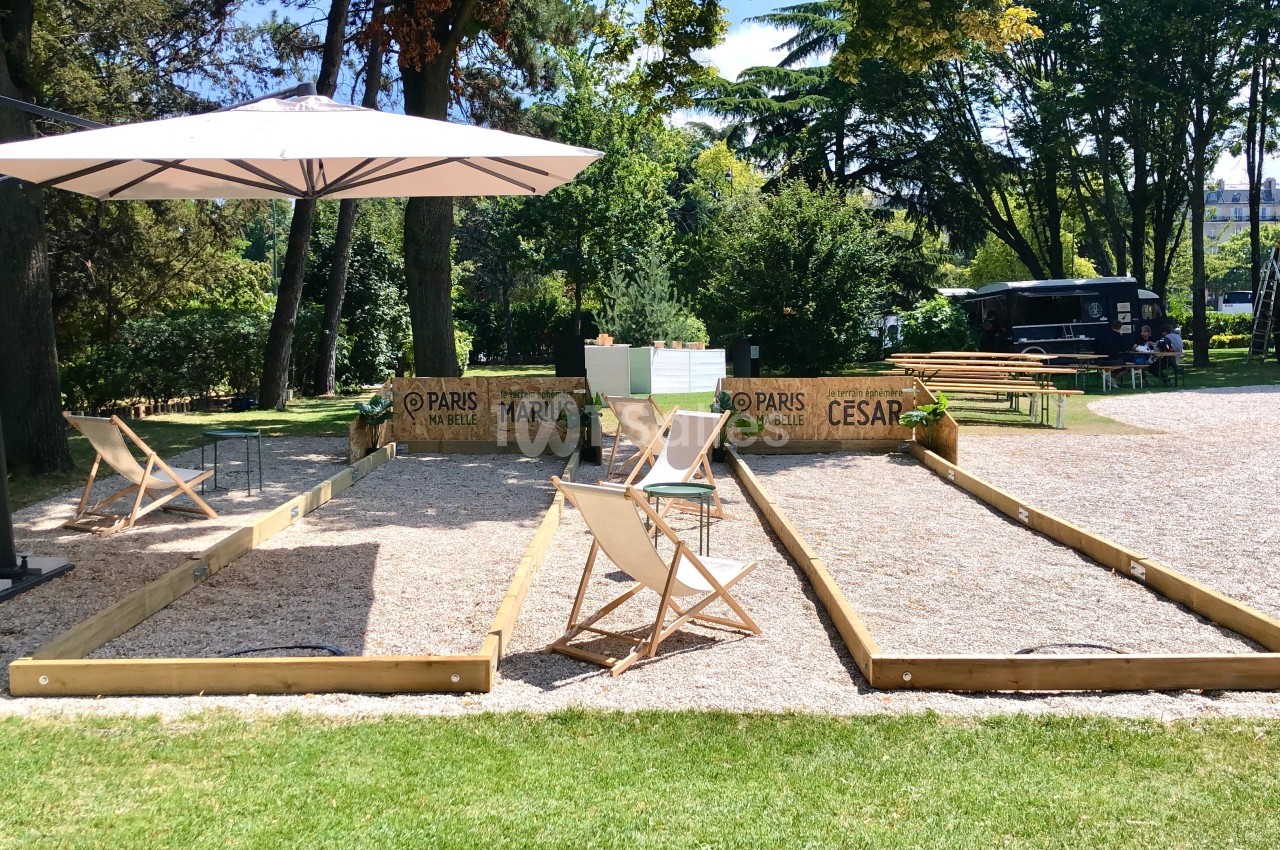 Terrain de pétanque en plein air avec chaises pliantes, parasol et espace ombragé entouré de verdure.