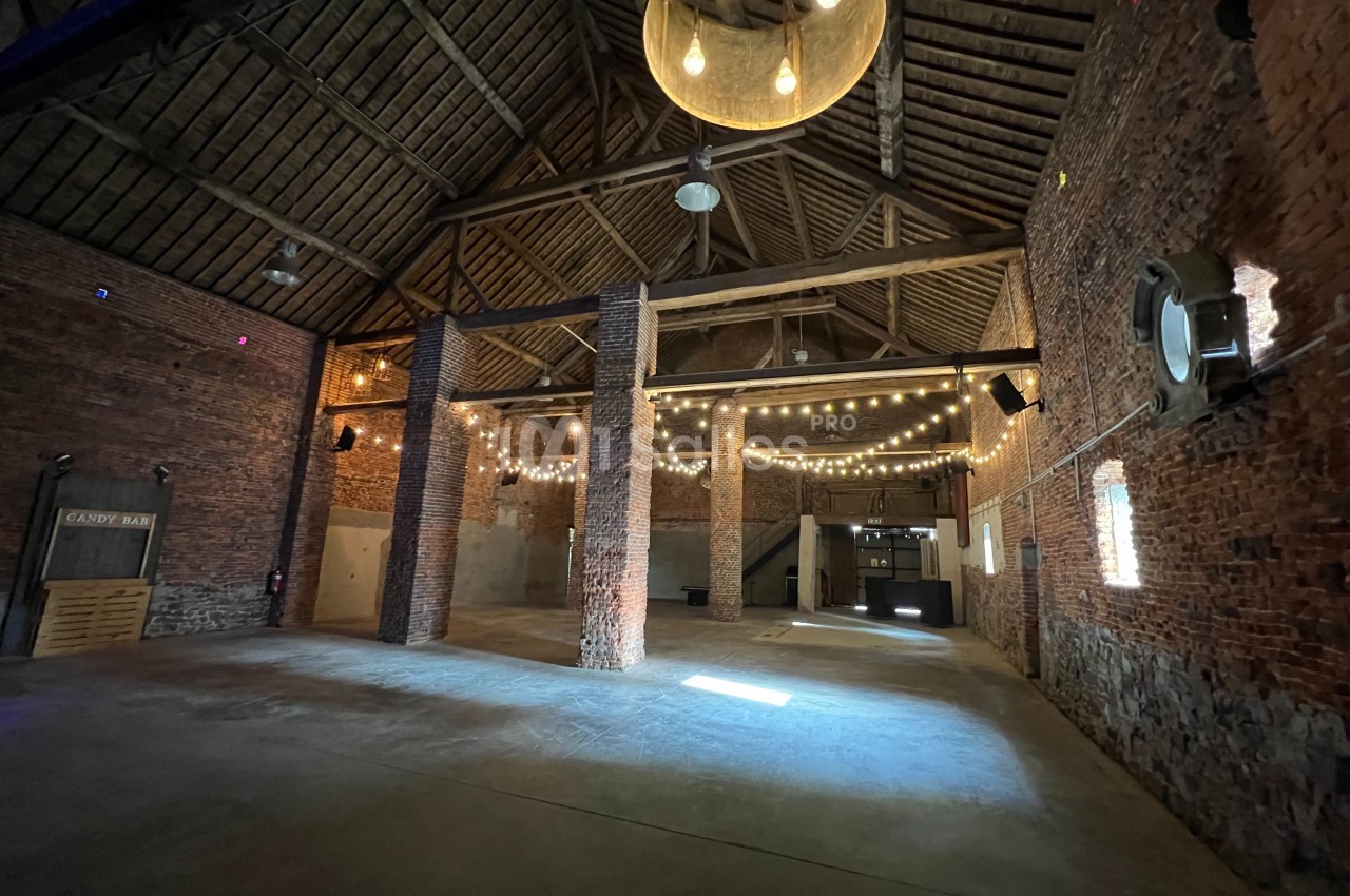 Intérieur d'une grande salle en briques avec charpente en bois, éclairée par des guirlandes lumineuses suspendues.