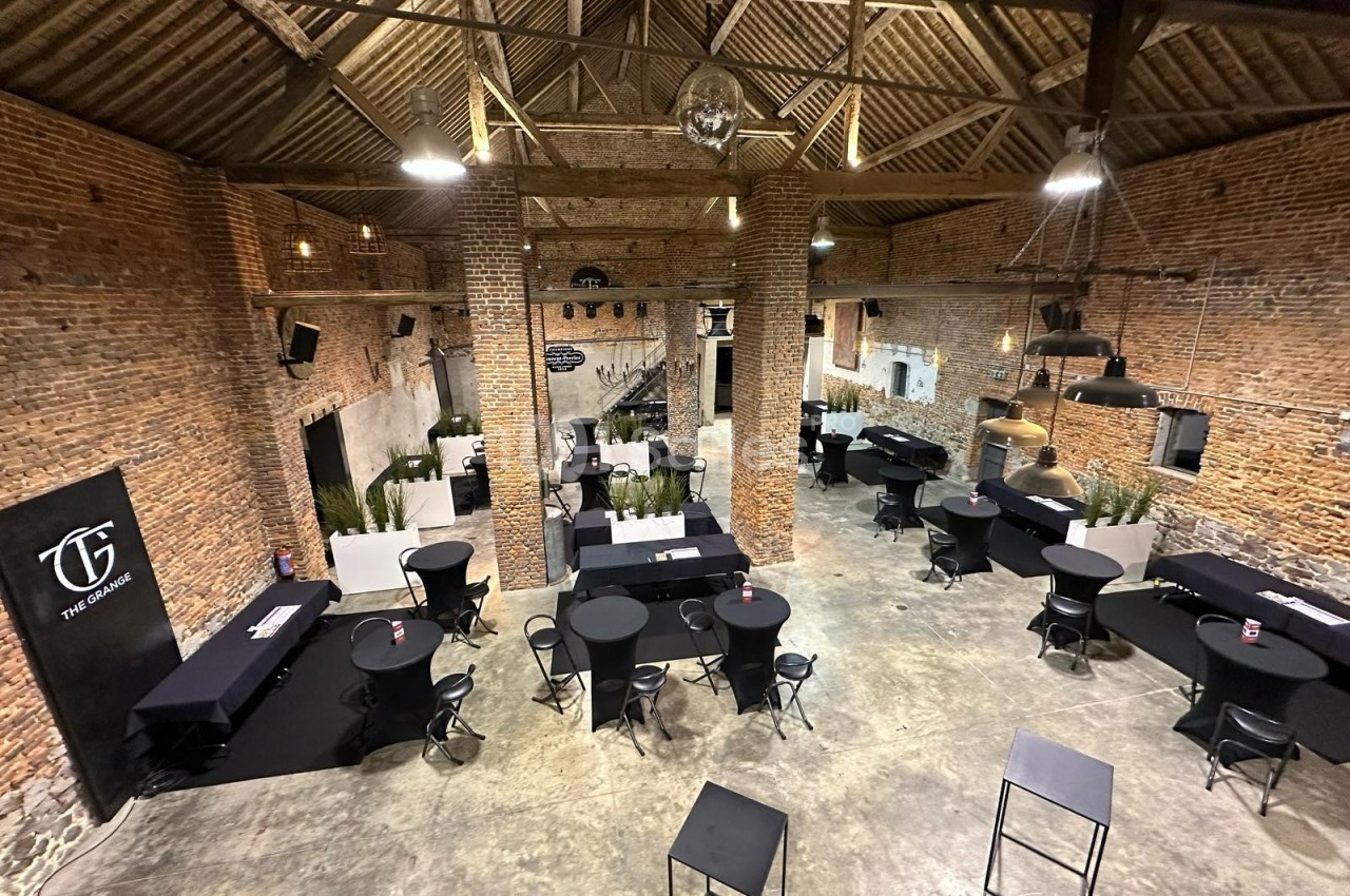 Salle en briques avec charpente apparente, aménagée avec des tables hautes et des chaises pour un événement.
