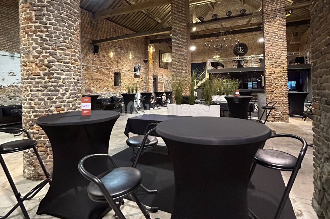 Salle en briques avec tables hautes noires et chaises, décorée de plantes et éclairée par des lustres suspendus.