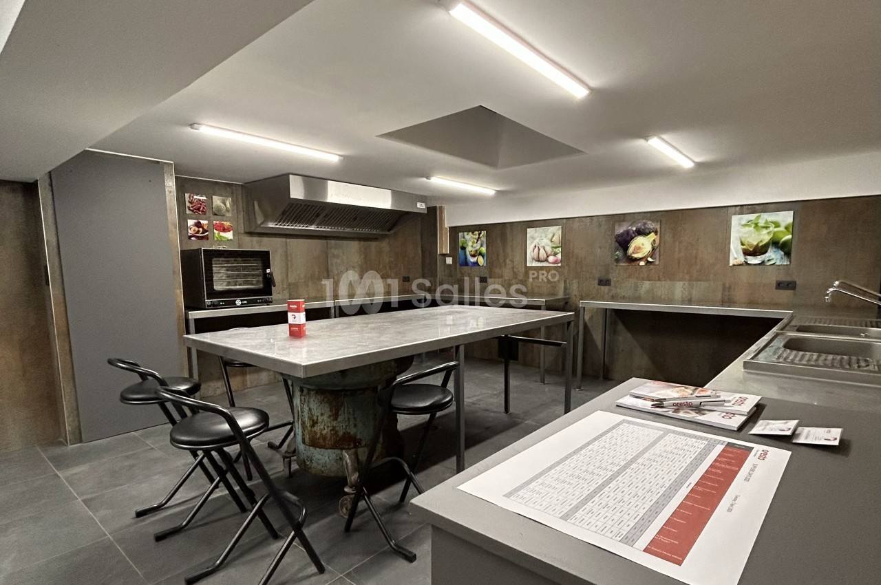 Cuisine professionnelle avec plans de travail en inox, tabourets, four encastré et affiches décoratives au mur.