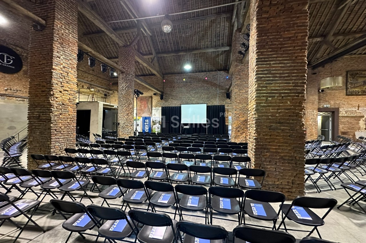Salle en briques avec des rangées de chaises noires alignées face à une scène équipée d'un écran de projection.