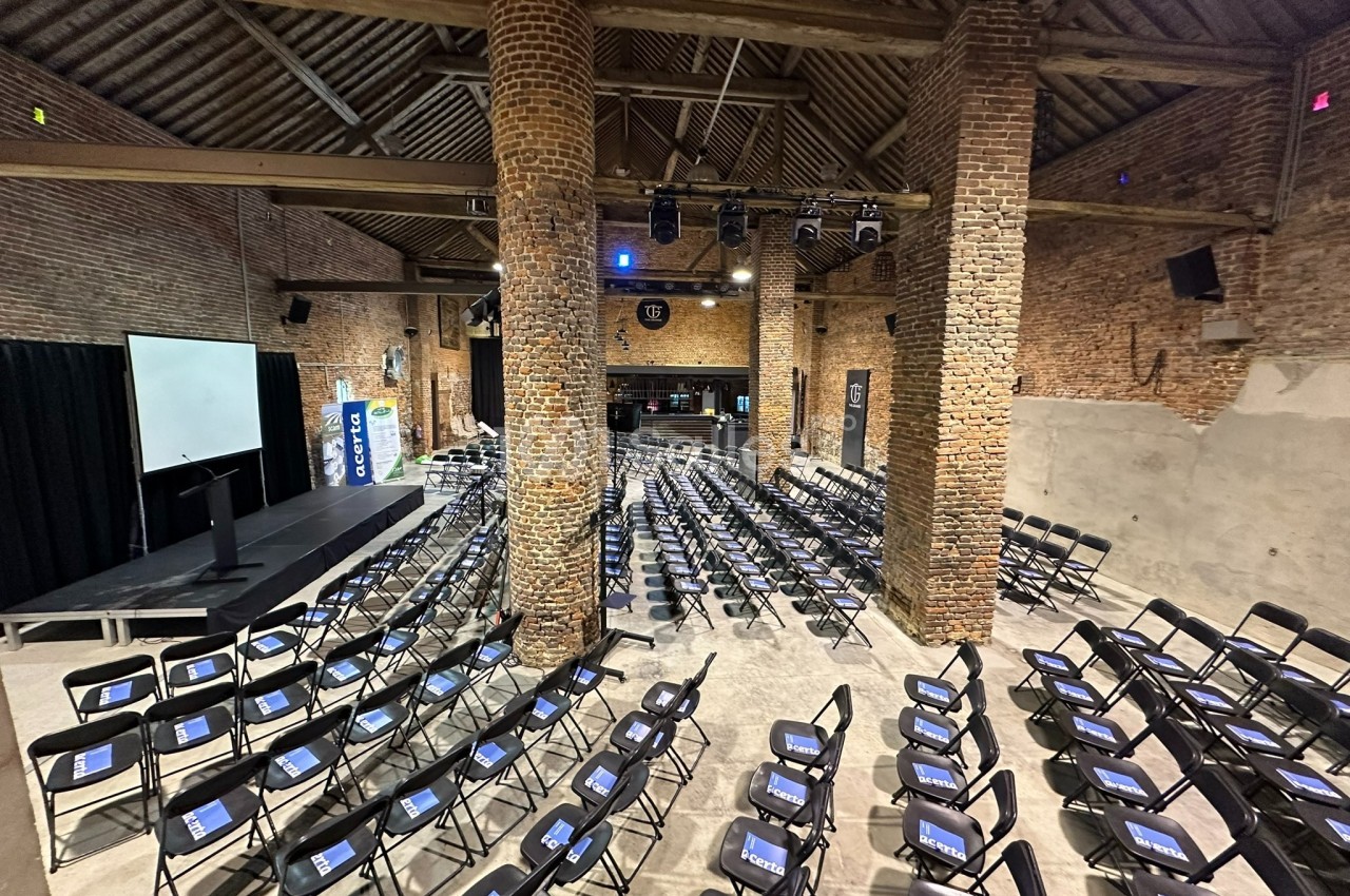 Salle en briques avec des rangées de chaises noires alignées, écran de projection et estrade à l'avant.