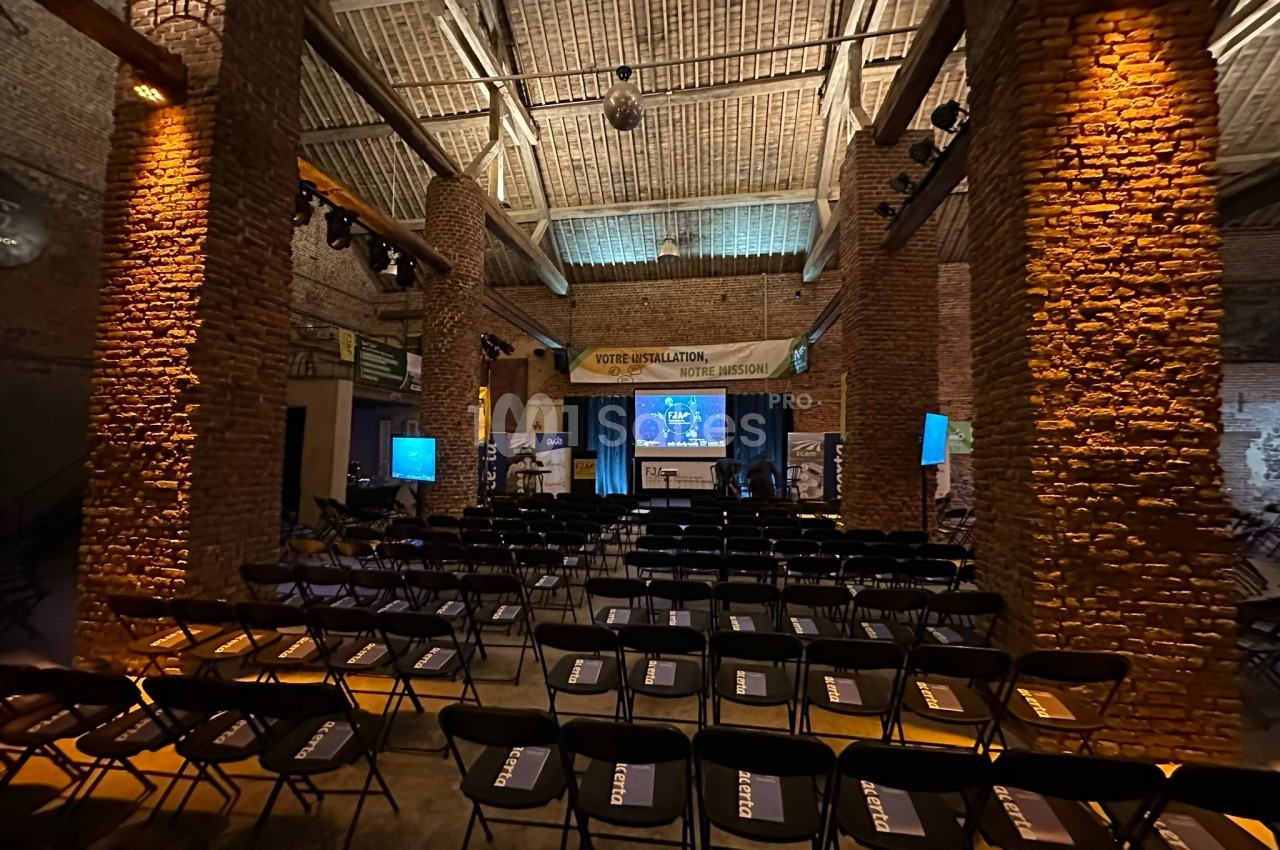 Salle de conférence en briques avec des rangées de chaises noires vides, un écran et une scène éclairée à l'arrière-plan.