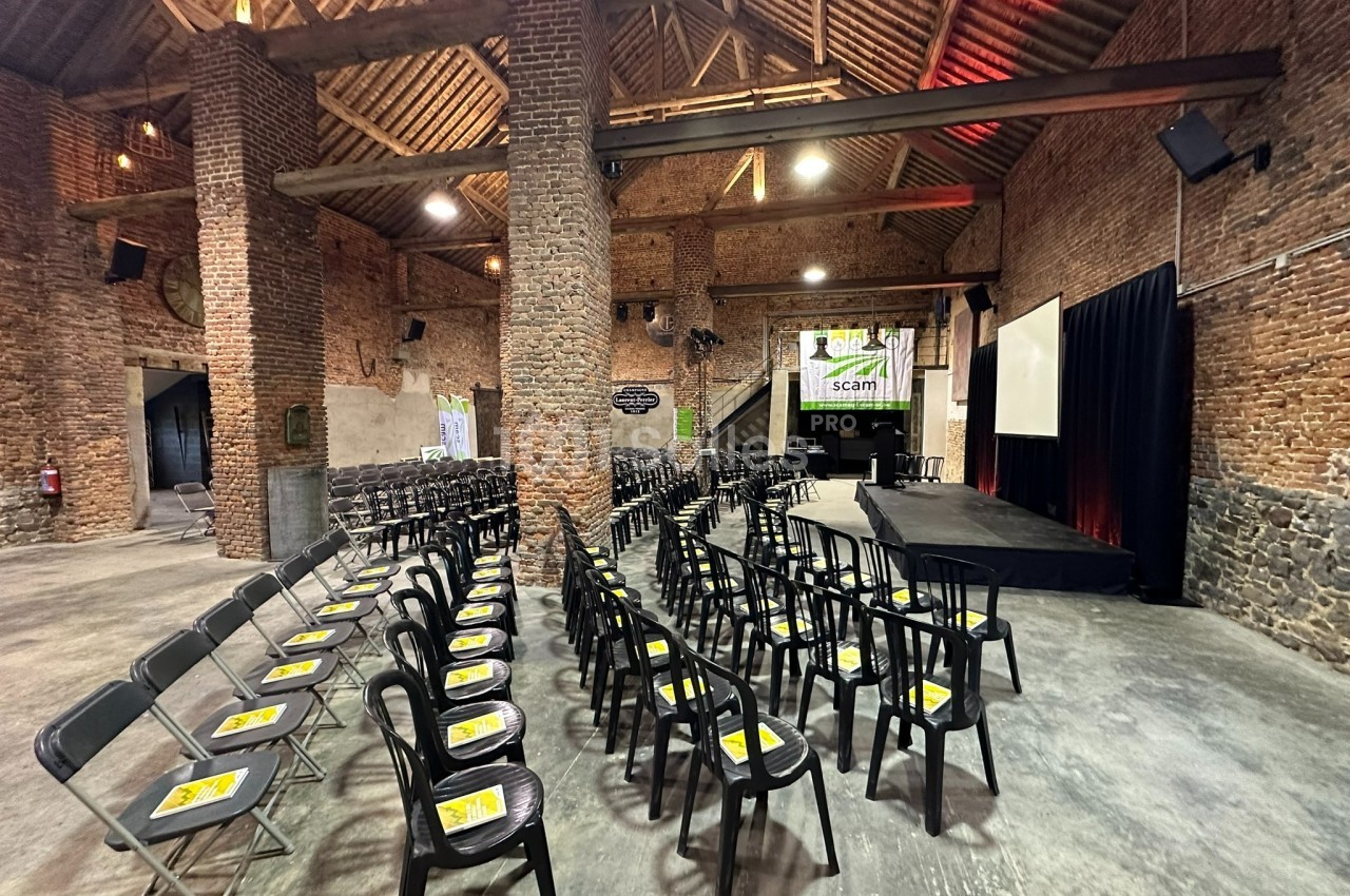Salle en briques avec des rangées de chaises noires alignées face à une scène et un écran de projection.